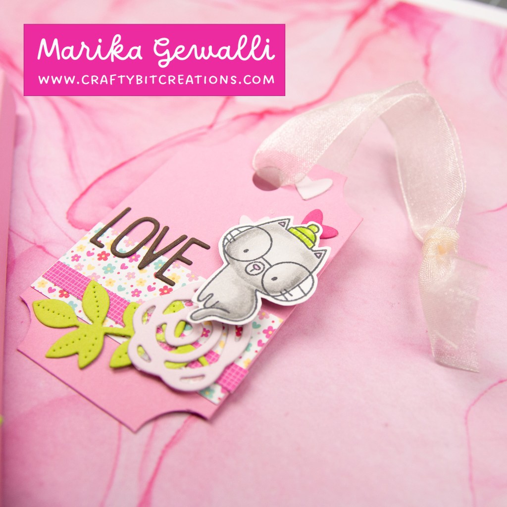 close up of mid pink tag, pattern papers with a diecut branch, rose and the letters spelling out love on top finally a stamped and colored cat on top of it all