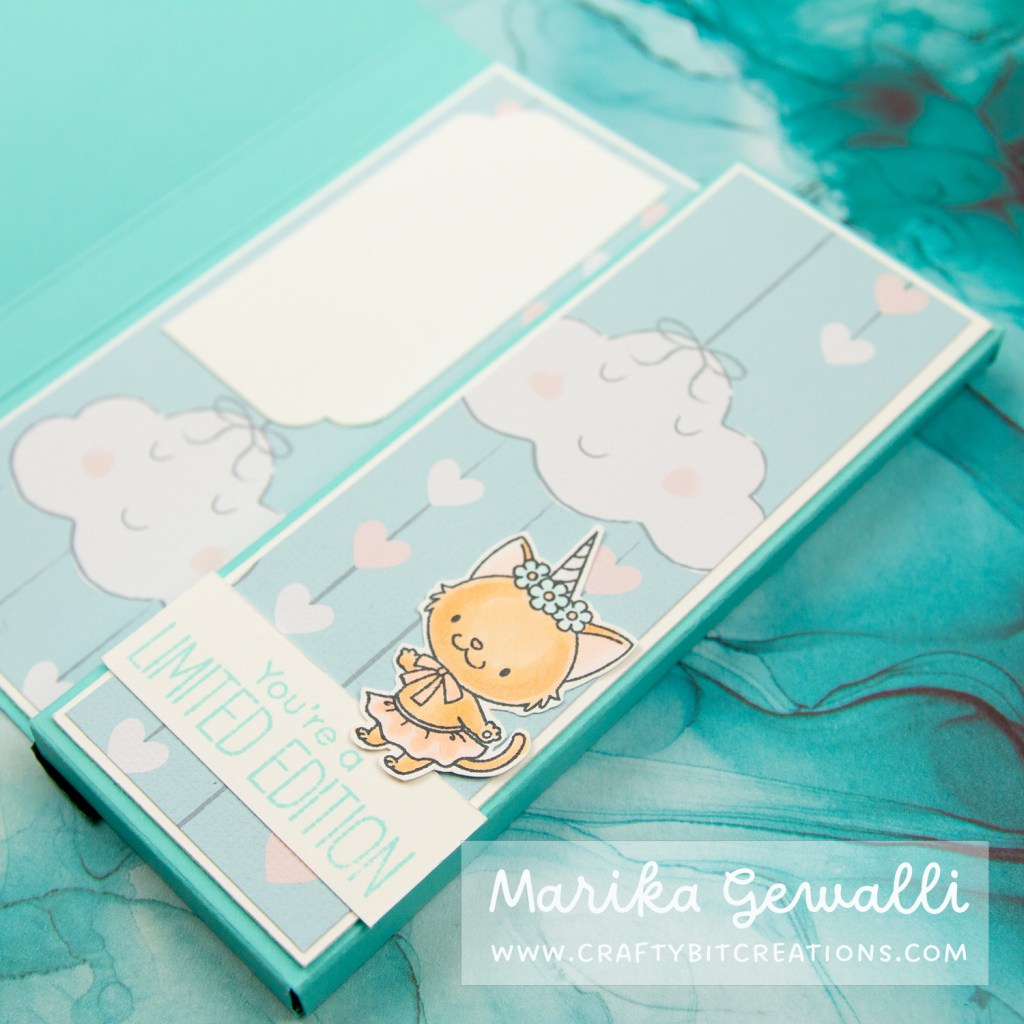 Close up of inside of a blue-green box card covered in pattern paper with clouds and strings of hearts. The closeup is focused on the unicorn cat with a skirt and wings that sits on top of a sentiment saying "you're a limited edition"