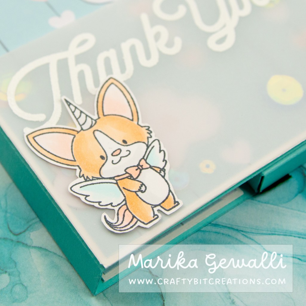 Close up image of a stamped cartoon corgy with wings and a unicorn horn, the colored image sits in the shaker part of the blue-green box card covered in pattern paper with clouds and strings of hearts.