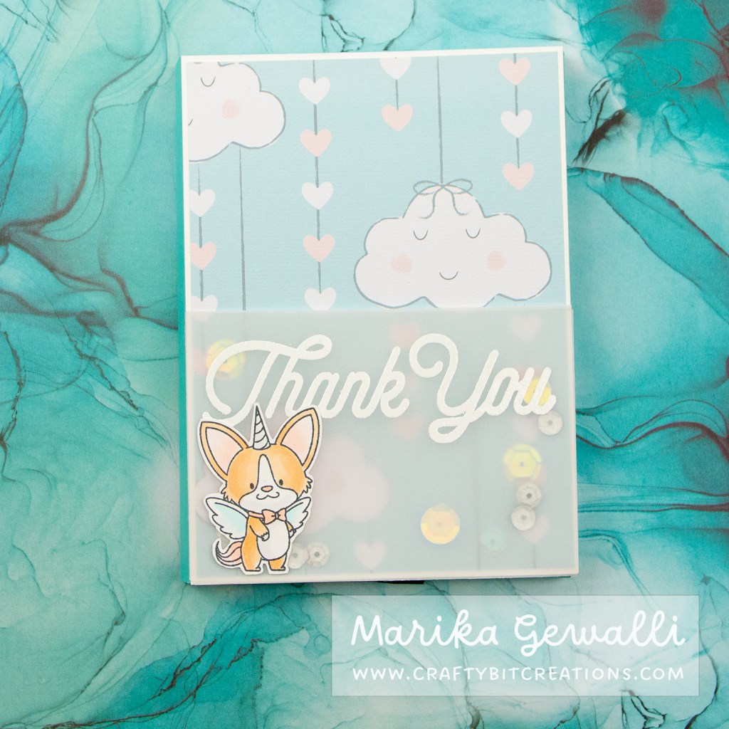 Close up images of a blue-green box card covered in pattern paper with clouds and strings of hearts. With a vellum shaker at the bottom decorated with a heat embossed sentiment saying "thank you" and a winged unicorn corgi with a bow.