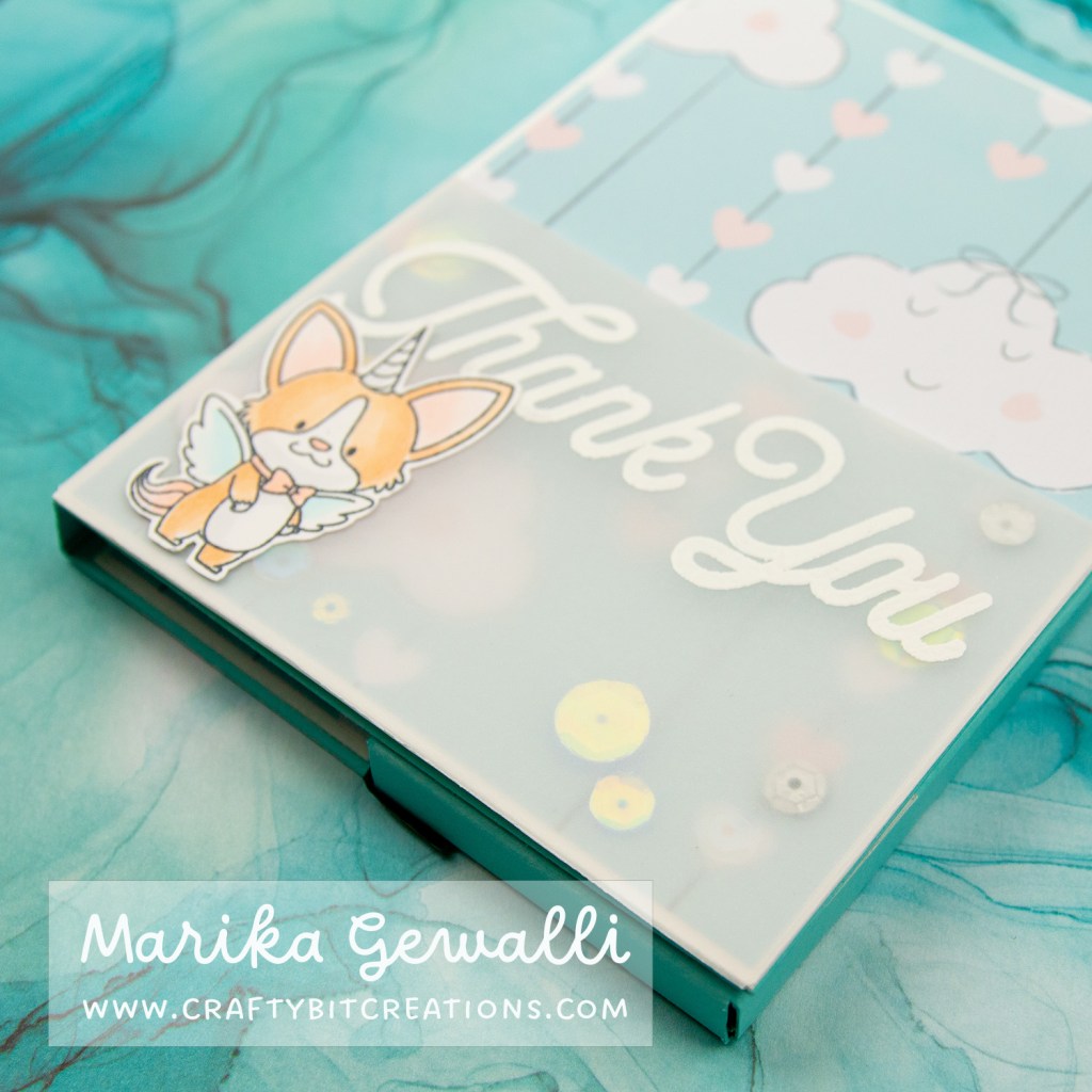 Close up images of a blue-green box card covered in pattern paper with clouds and strings of hearts, on the lower part it has a shaker made out of vellum with a Thank you stamped and heat embossed in white on the top and a winged unicorn corgi at the bottom left corner.