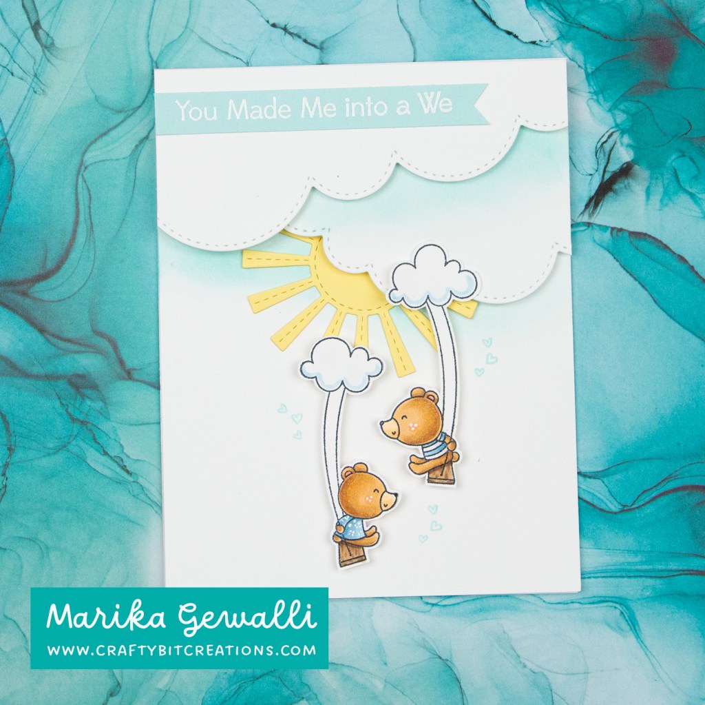 Picture of card with clouds on top with a sentiment strip in sno cone cardstock with white embossed sentiment "you made me into a we". under the clouds is a sun and two bears swinging from clouds. Around them are mini hearts stamped in sno cone ink